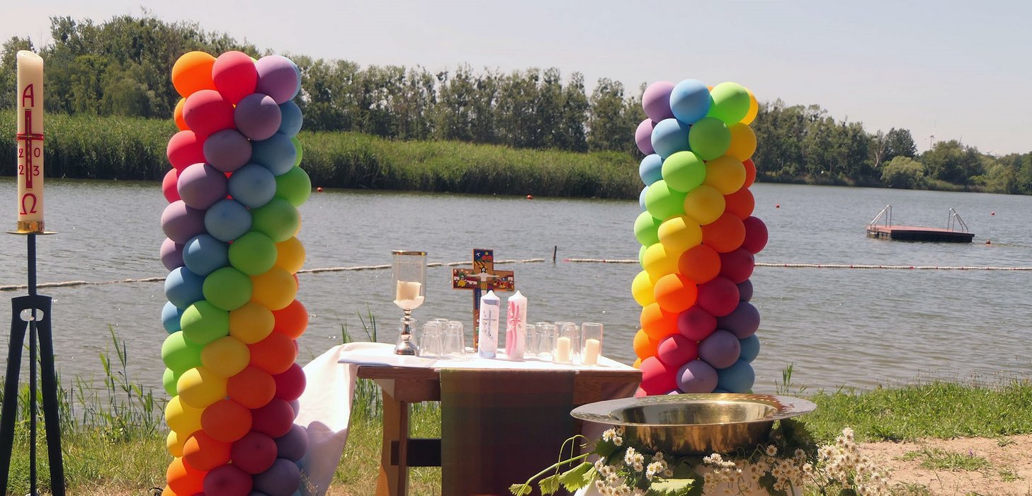 Ein Tisch mit Kerzen, Gläsern und einem Kreuz steht für eine Taufe am Ufer eines Sees. Daneben zwei regenbogenfarbene  Luftballon Türme und im Vordergrund eine Taufschale.