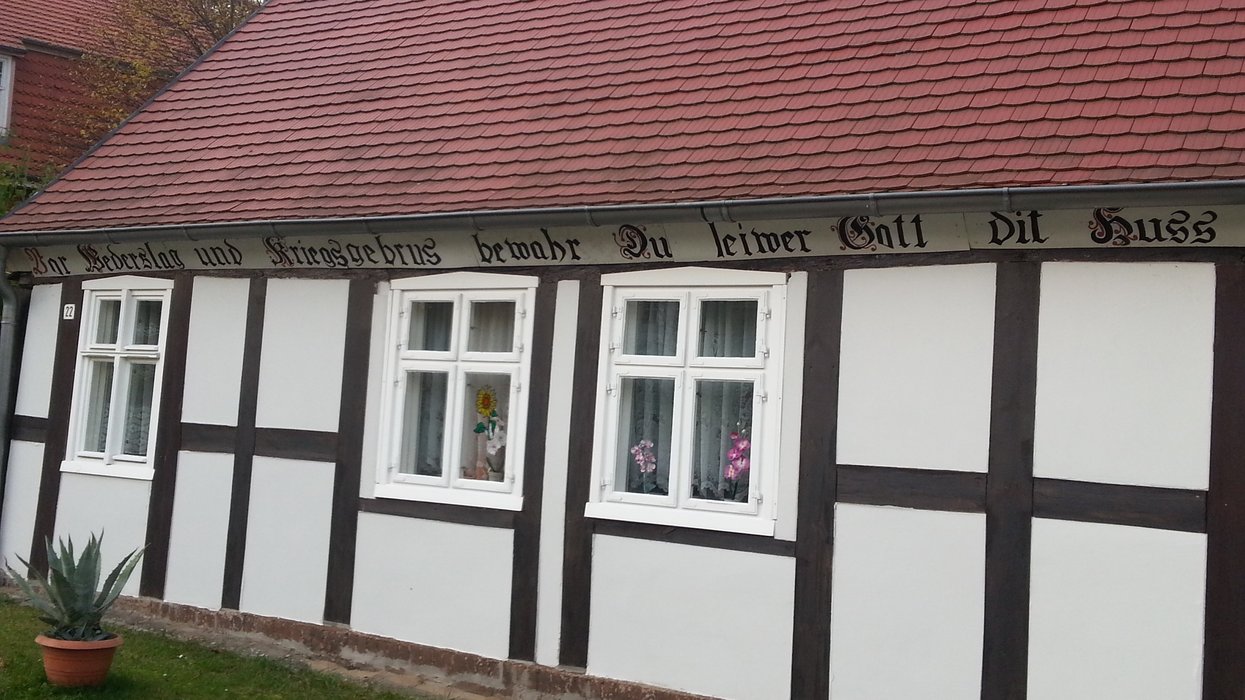 Weiße Hauswand mit Holzbalken und drei Fenstern, auf der ein plattdeutscher Segensspruch steht.