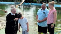 Ein Junge wird in einem See getauft. Daneben stehen zwei Erwachsene und im Hintergrund ist ein Wasserspielplatz.