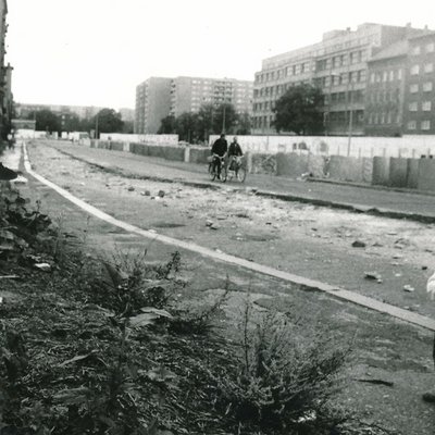 Schwarz-Weiß-Bild der Berliner Mauer. Zu sehen ist ein Kind mit Hammer vor der Mauer, eine Sonnenblume, im Hintergrund zwei Radfahrer.