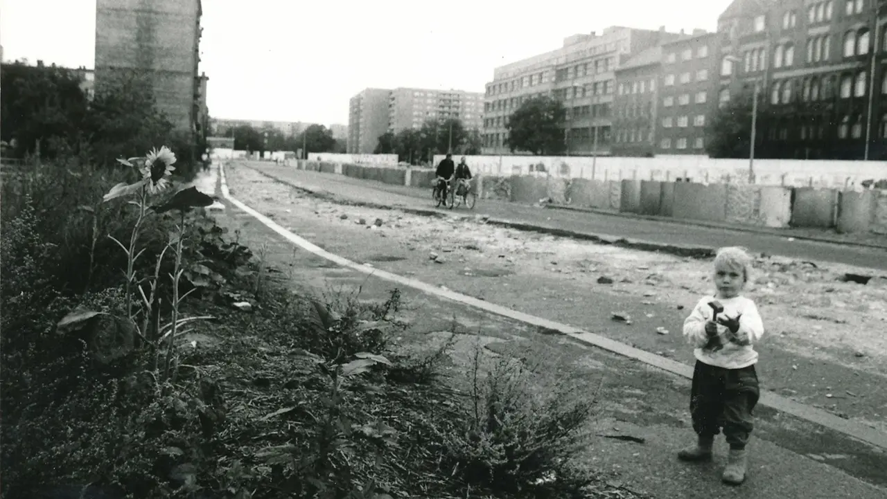 Schwarz-Weiß-Bild der Berliner Mauer. Zu sehen ist ein Kind mit Hammer vor der Mauer, eine Sonnenblume, im Hintergrund zwei Radfahrer.