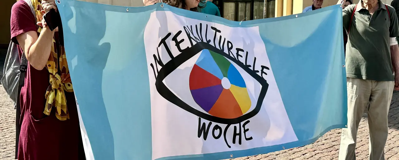 Das Logo der interkulturellen Woche eines menschlichen Auges mit einer mehrfarbigen Iris in Tortendiagrammform wird von Menschen auf einem Transparent hochgehoben.