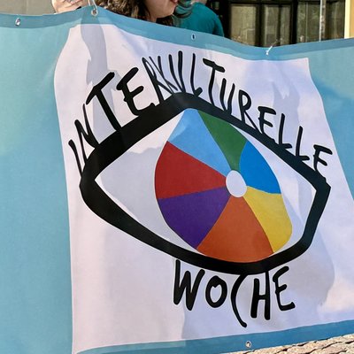 Das Logo der interkulturellen Woche eines menschlichen Auges mit einer mehrfarbigen Iris in Tortendiagrammform wird von Menschen auf einem Transparent hochgehoben.