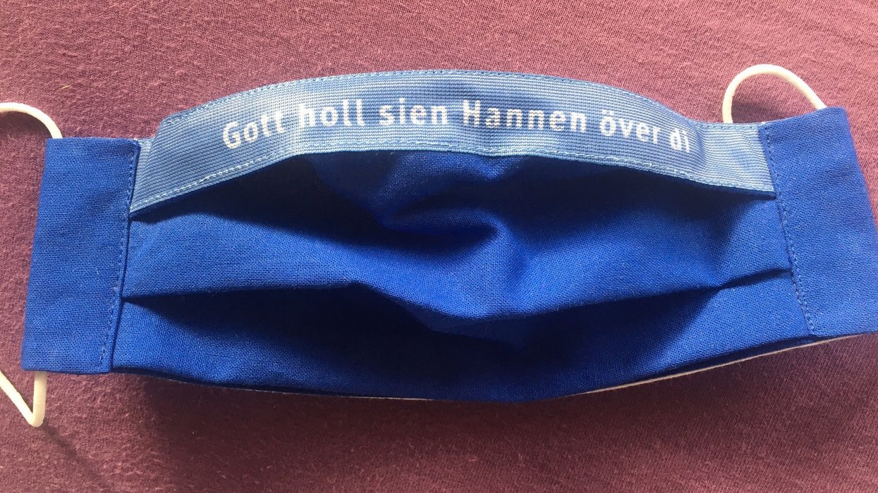 Blaue Stoffmaske mit der Aufschrift ,,Gott holl sien Hannen över di''.