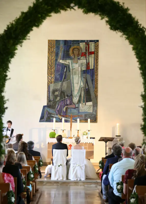 hepaar sitzt auf Stühlen vor dem Altarraum. Braut in weißem Kleid. Bräutigam links daneben in schwarzem Anzug. Links sieht man die Band.