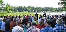 Eine Gruppe von Menschen sitzt an einem See auf Bänken in Blickrichtung zu zwei Pfarrern, die zu den Menschen sprechen.