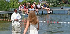 Ein Mädchen im weißen Kleid wird von einer Frau im weißen Gewand in einem See getauft. Am Rand sitzen Menschen auf einem Steg und schauen zu.